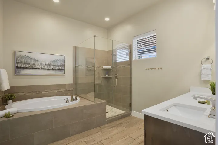 Built-in soaking tub, glass-enclosed shower with tiled walls and floor, dual vanity with undermount sinks, and wood-finish flooring