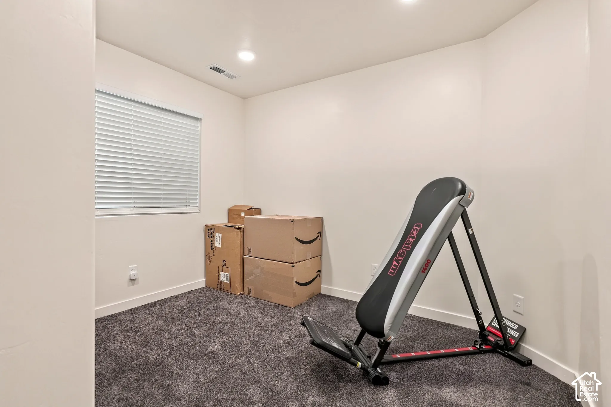 Recessed lighting illuminates the room, featuring a window with white blinds and dark gray carpeting