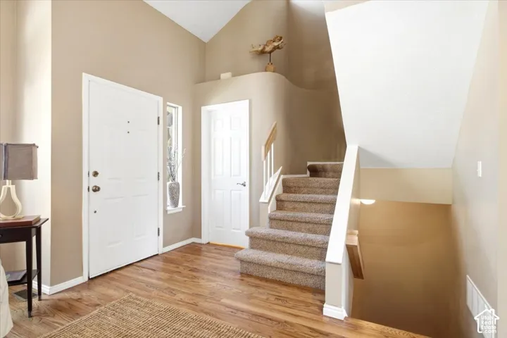 Vaulted ceiling entry with wood-finish flooring, a white front door with sidelight, and a carpeted staircase featuring a wood handrail