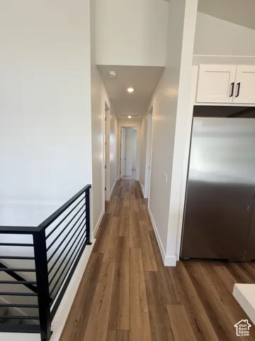 Wood-finish flooring extending through a hallway, featuring recessed lighting, white cabinetry with black hardware, and a stainless steel refrigerator