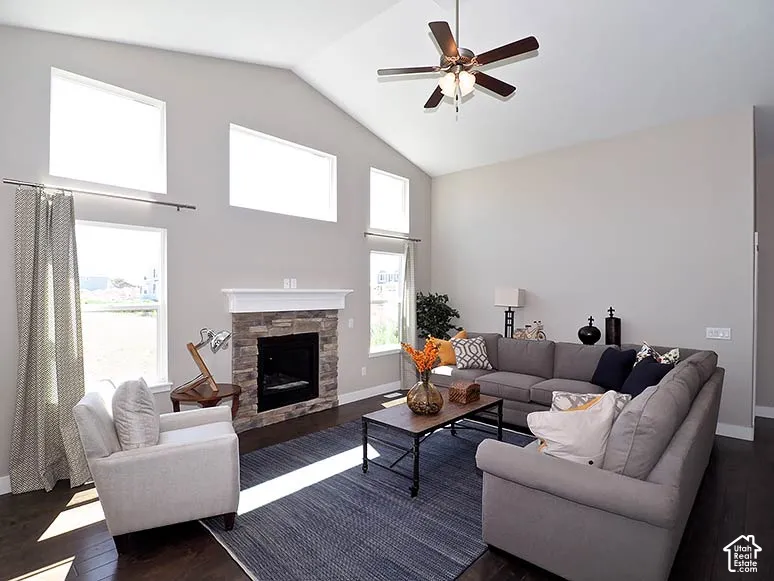Vaulted ceiling with a ceiling fan, stone-faced fireplace with a white mantel, multiple large windows, and dark wood-finish flooring
