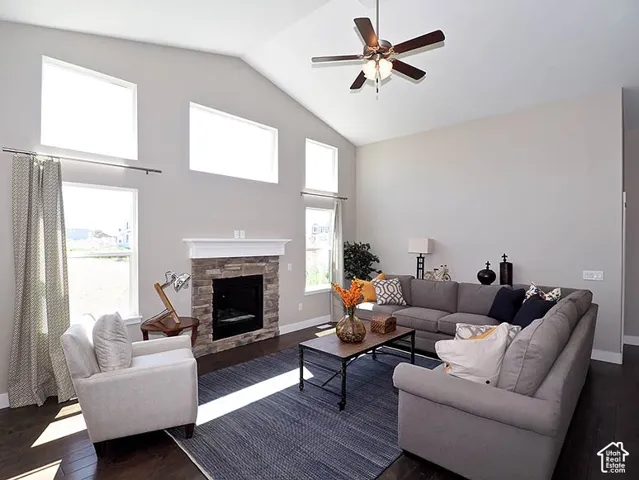 Vaulted ceiling with a ceiling fan, stone-faced fireplace with a white mantel, multiple large windows, and dark wood-finish flooring