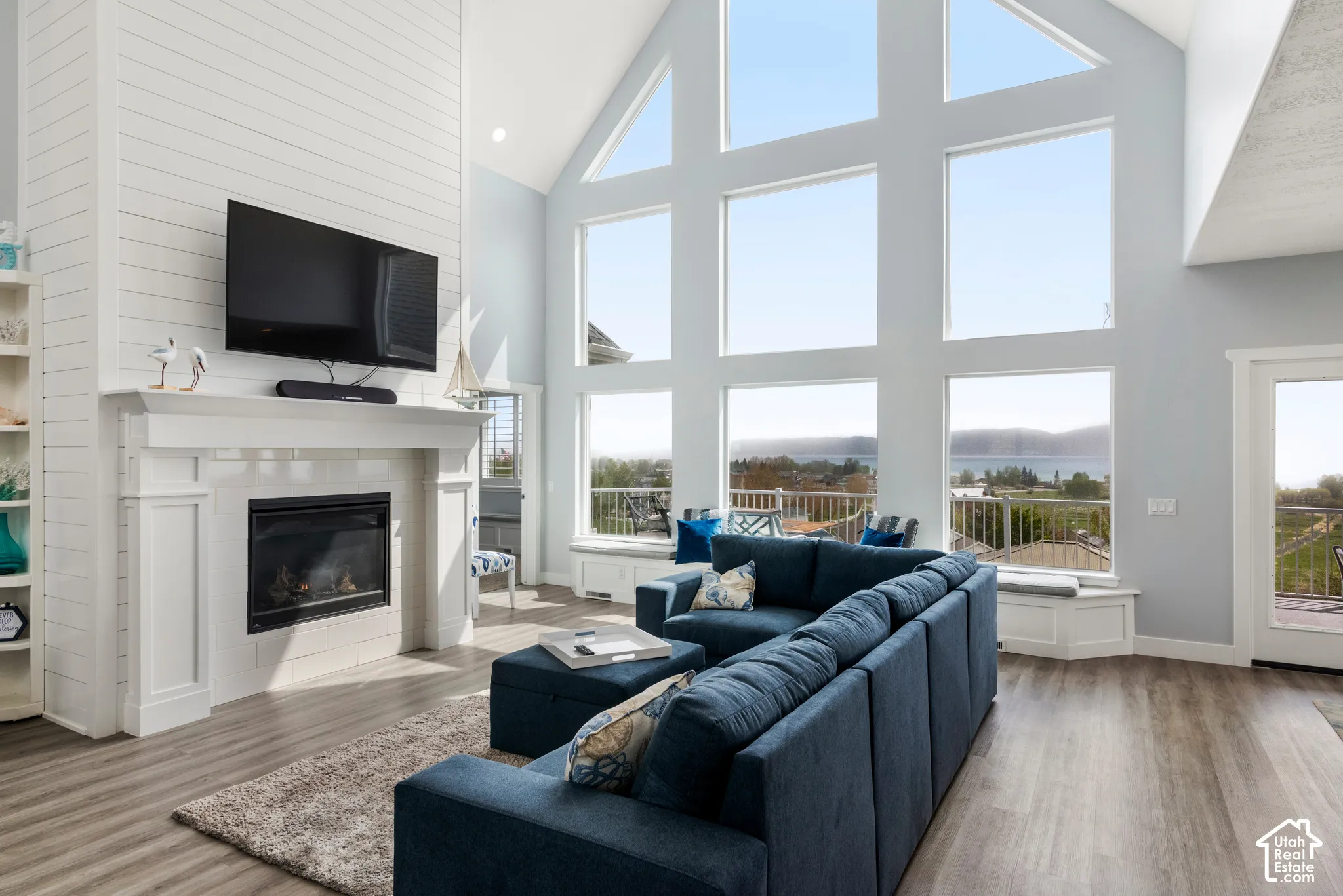 Vaulted ceiling, shiplap-paneled fireplace with mantel, large picture windows, wood-finish flooring, and built-in window seating