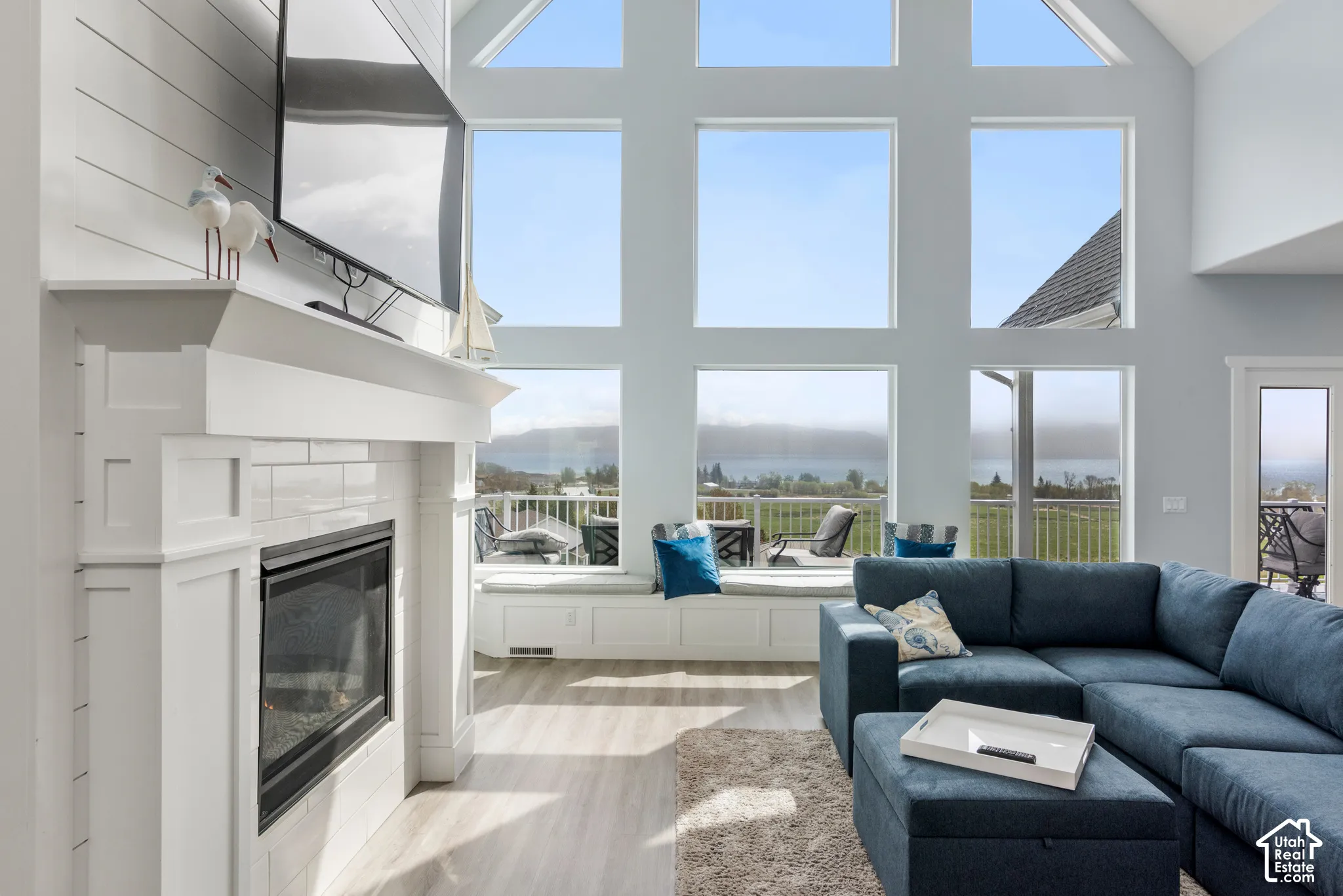 Vaulted ceiling with extensive window array, shiplap-accented fireplace with white tile surround, built-in window seating, and wood-finish flooring
