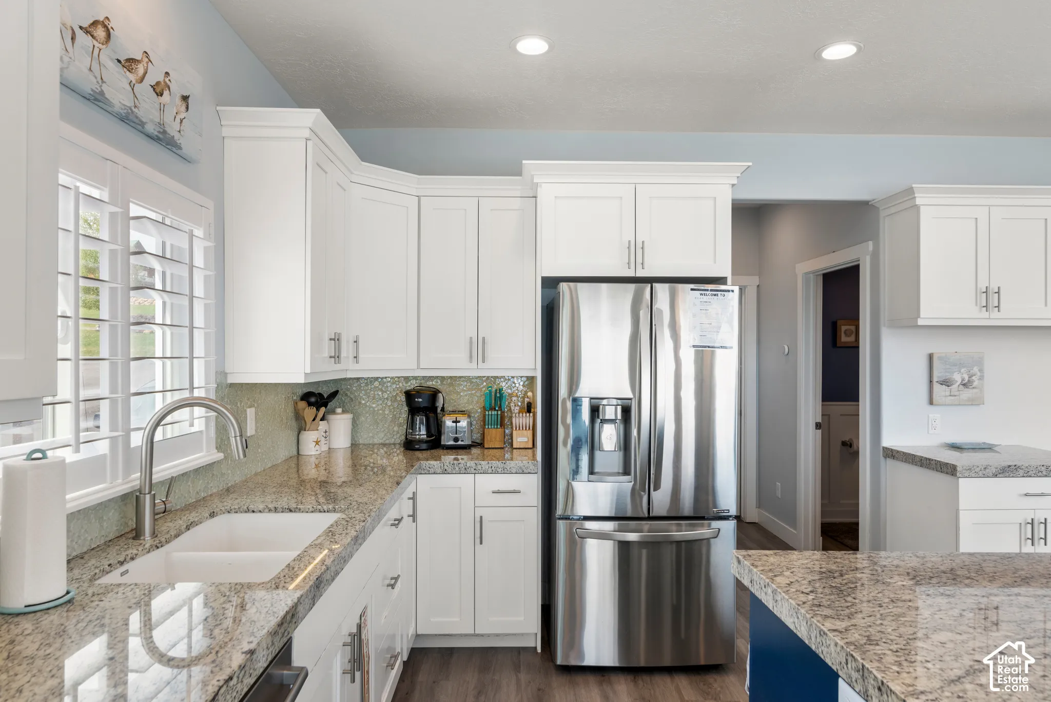 White shaker-style cabinetry with crown molding, granite countertops, a built-in stainless steel refrigerator, a double basin sink with a gooseneck faucet, and wood-finish flooring