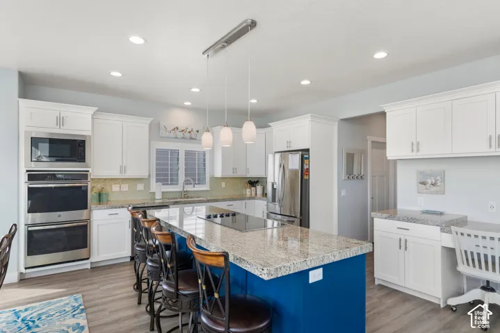 Kitchen featuring white cabinetry, wood-finish flooring, stainless steel appliances, a large center island with a cooktop, and a built-in desk area