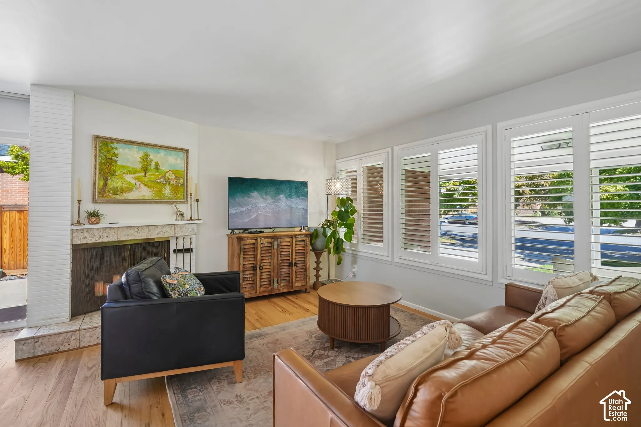 Hardwood flooring, white painted brick fireplace with stone surround, multiple windows with white plantation shutters, and recessed lighting