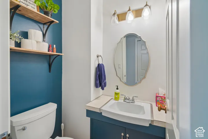 Vanity with integrated sink, gold-toned wall-mounted light fixture, decorative mirror, dual-shelf built-in storage, and white commode