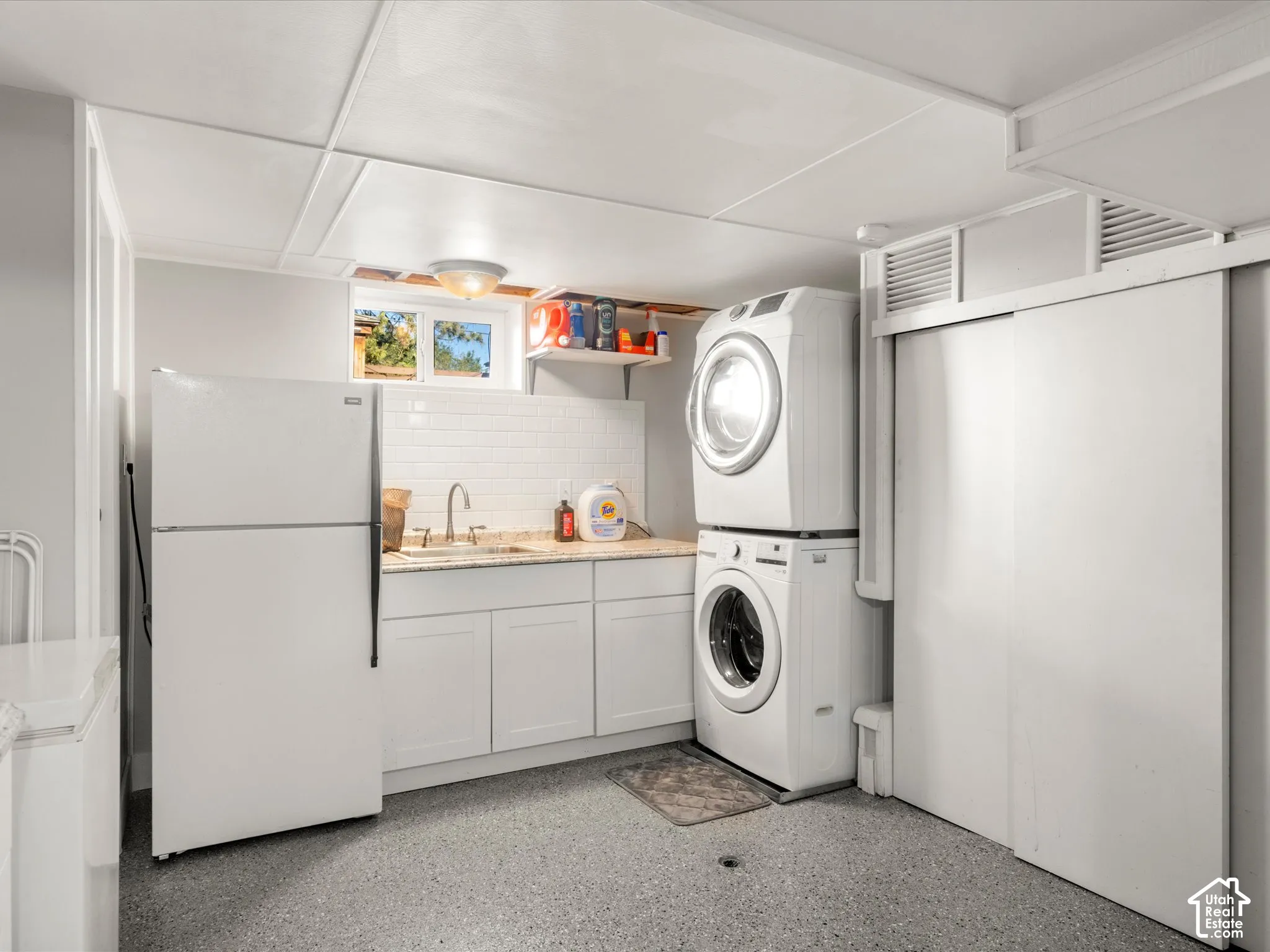 Laundry area featuring white stacked washer and dryer, white cabinetry, a sink with a chrome faucet, and a subway tile backsplash