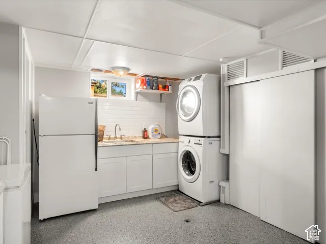 Laundry area featuring white stacked washer and dryer, white cabinetry, a sink with a chrome faucet, and a subway tile backsplash
