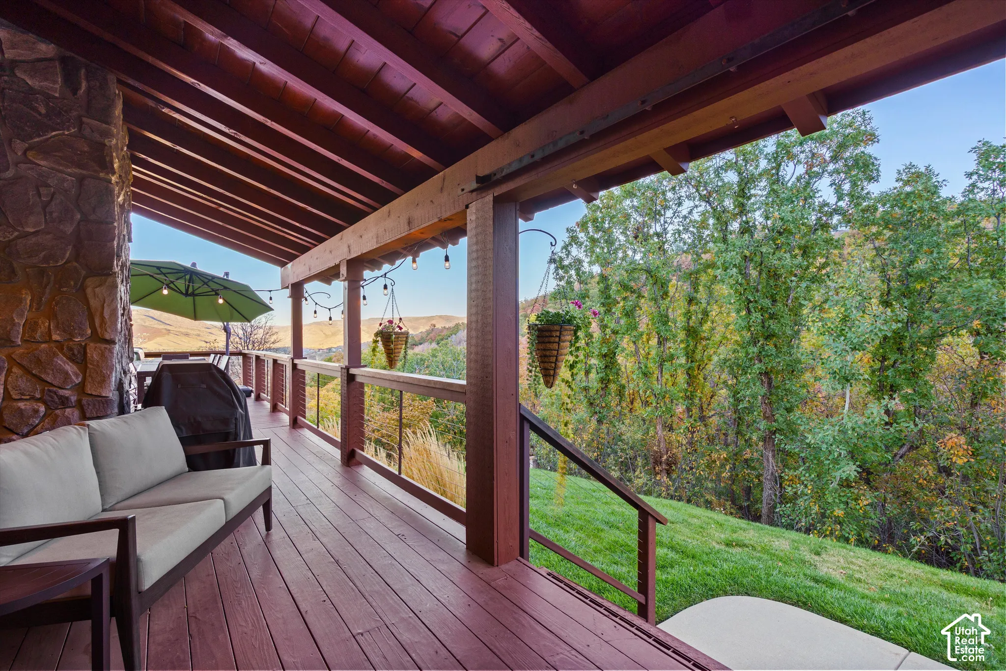 Expansive covered deck with natural wood flooring and exposed rafters, featuring a stone-clad exterior wall and a clear railing system overlooking a wooded hillside