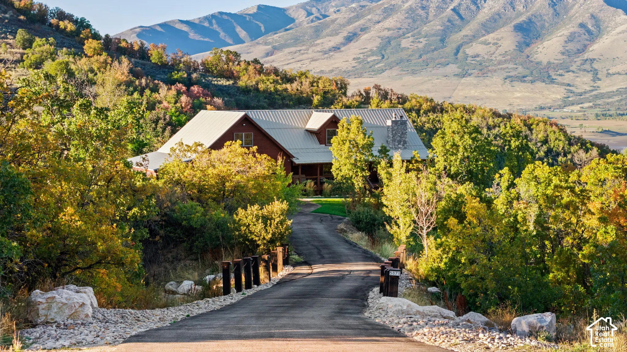 Paved driveway leading to a residence with a metal roof, stone chimney, and wood siding, set amidst mature trees with a mountain backdrop