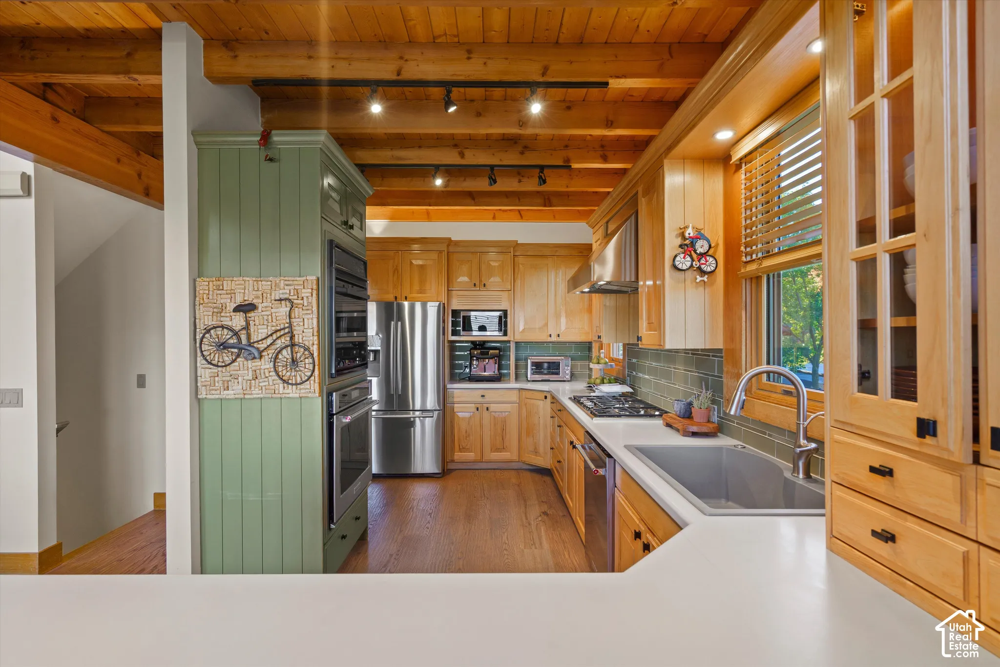 Exposed wood beam ceiling, wood-finish flooring, built-in stainless steel appliances, light wood cabinetry with glass-front uppers, and a tile backsplash