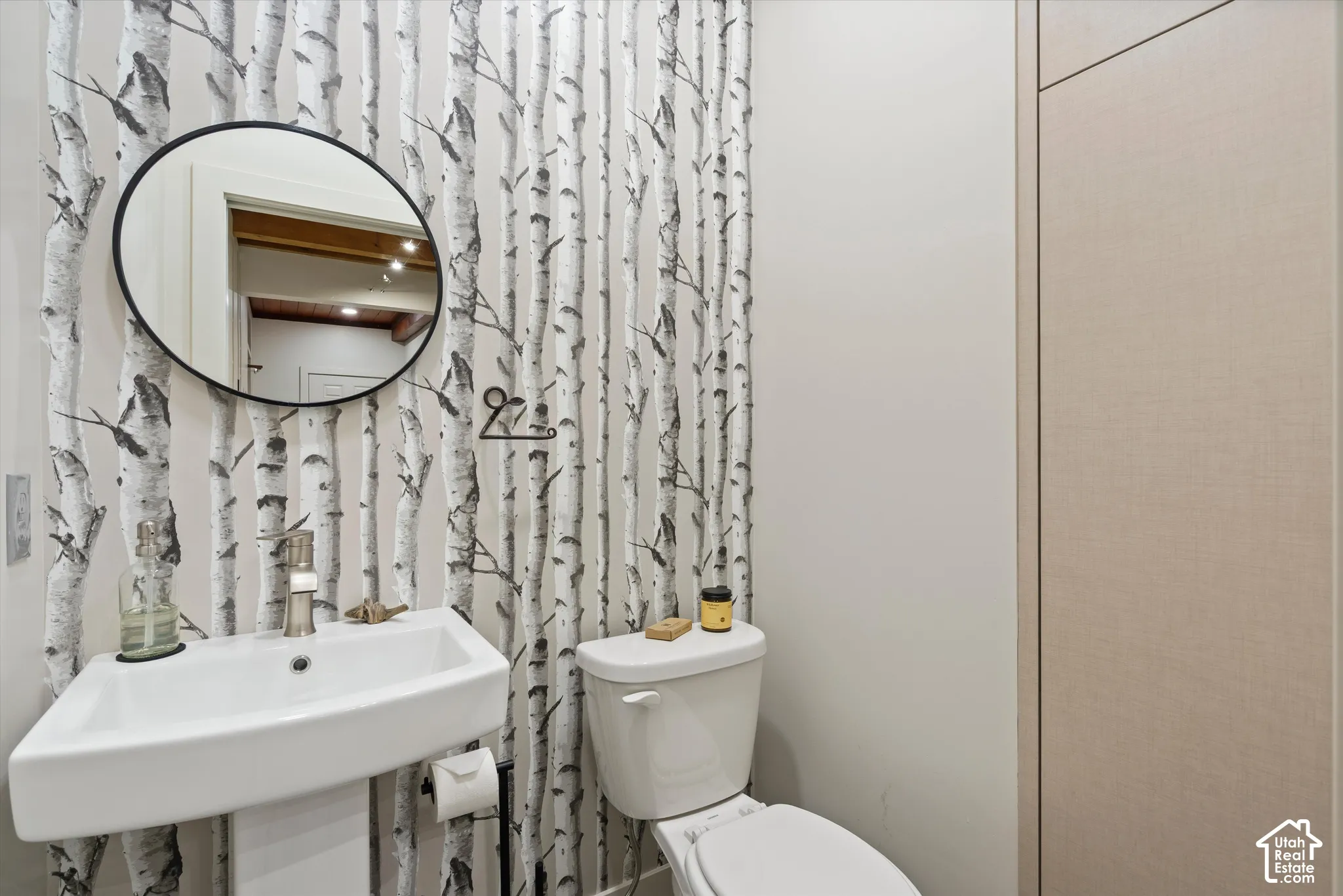 Pedestal sink with a wall-mounted round mirror, birch-patterned accent wall, and integrated flush cabinetry