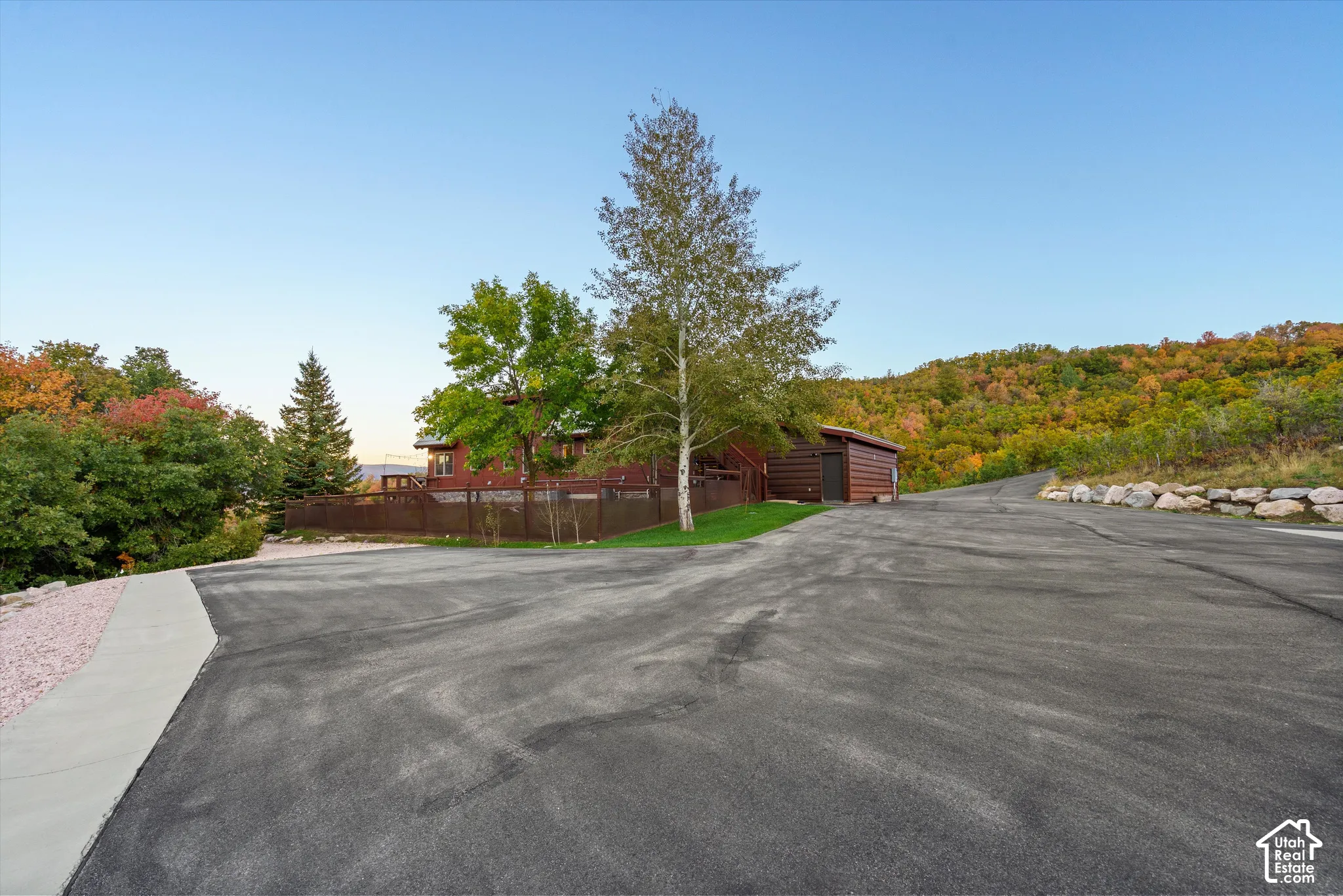 Asphalt driveway with an adjacent concrete walkway, leading to a log-style shed and a multi-level wood-sided residence, surrounded by mature trees and a natural rock retaining wall