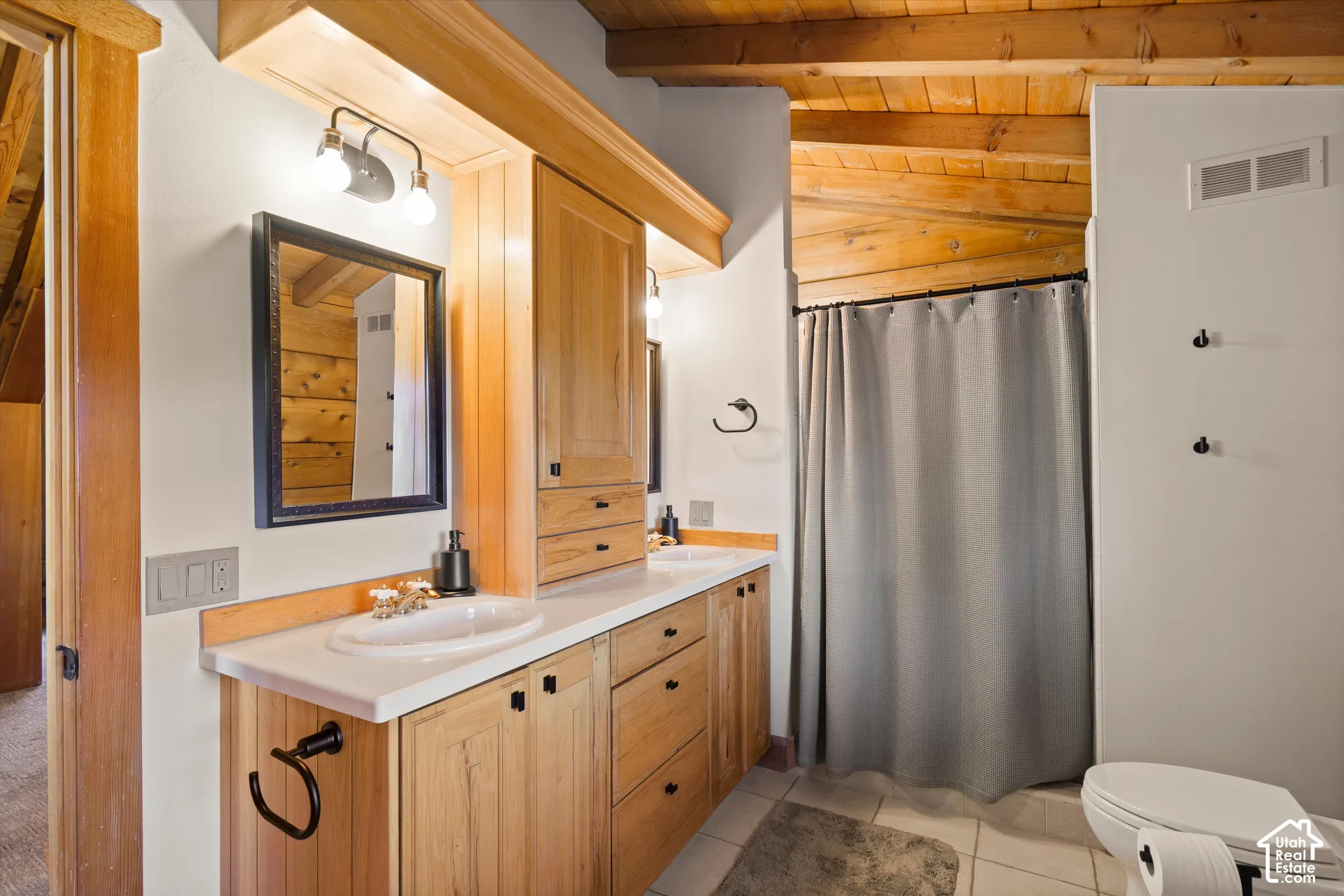 Vaulted wood-beam ceiling, dual vanity with undermount sinks, wood cabinetry, white countertops, and tile flooring