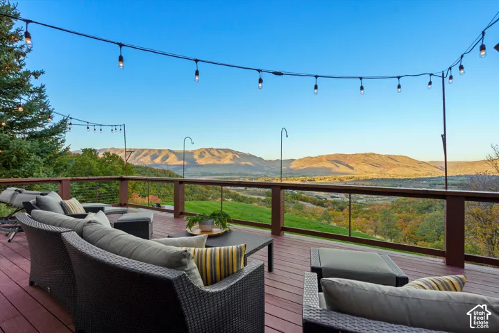 Expansive wood-finish deck with a glass-paneled railing, strung outdoor lighting, and a panoramic mountain backdrop