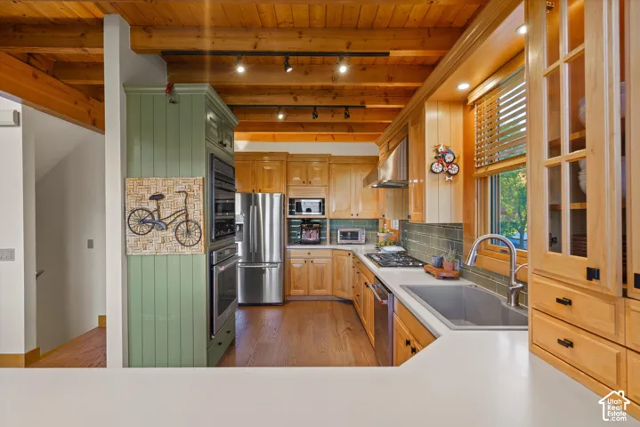 Exposed wood beam ceiling, wood-finish flooring, built-in stainless steel appliances, light wood cabinetry with glass-front uppers, and a tile backsplash
