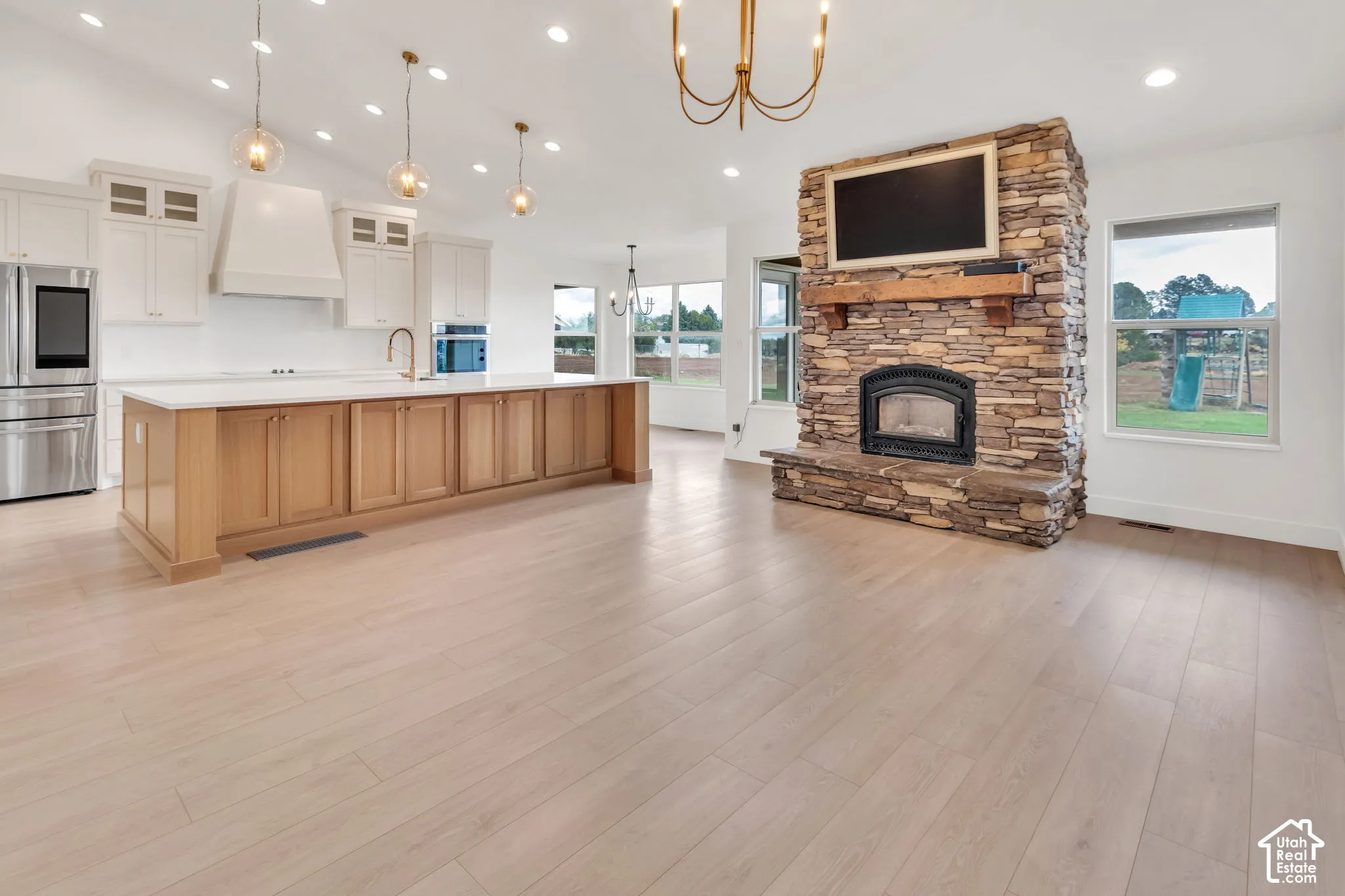 Open-concept living space with wood-finish flooring, a stone-clad fireplace with a wood mantle, and a kitchen island with light wood cabinetry