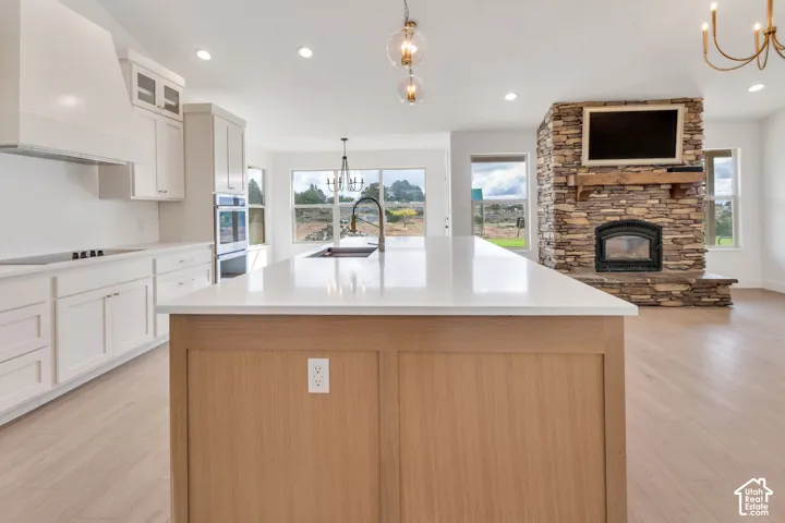 White shaker cabinetry with an integrated range hood, a kitchen island featuring a light wood-finish base, dual wall ovens, and a stacked stone fireplace with a wood-finish mantel and hearth