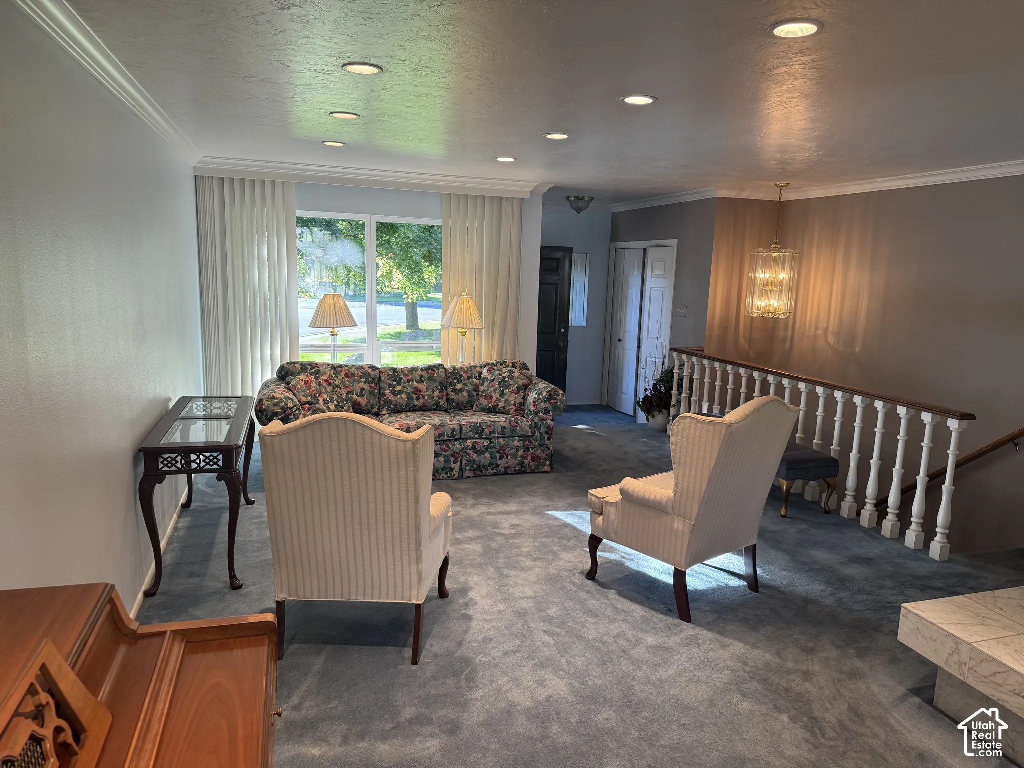 Recessed lighting throughout, crown molding, large picture window with drapes, staircase with wood handrail and white balusters, and a chandelier