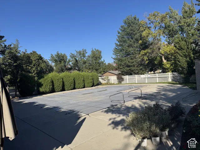 Expansive paved patio with a covered in-ground pool, surrounded by mature trees and a white privacy fence
