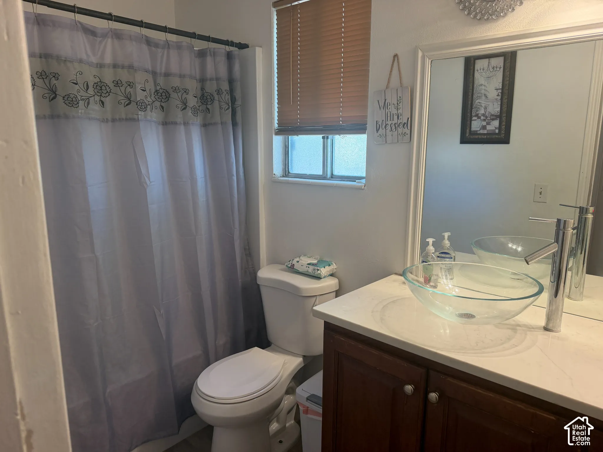 Bathroom featuring a glass vessel sink, chrome faucet, wood-finish vanity with white countertop, wall-mounted mirror, and a window with blinds