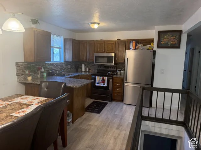 Kitchen featuring wood-finish cabinetry, granite-style countertops, stainless steel appliances, a tiled backsplash, and light wood-finish flooring