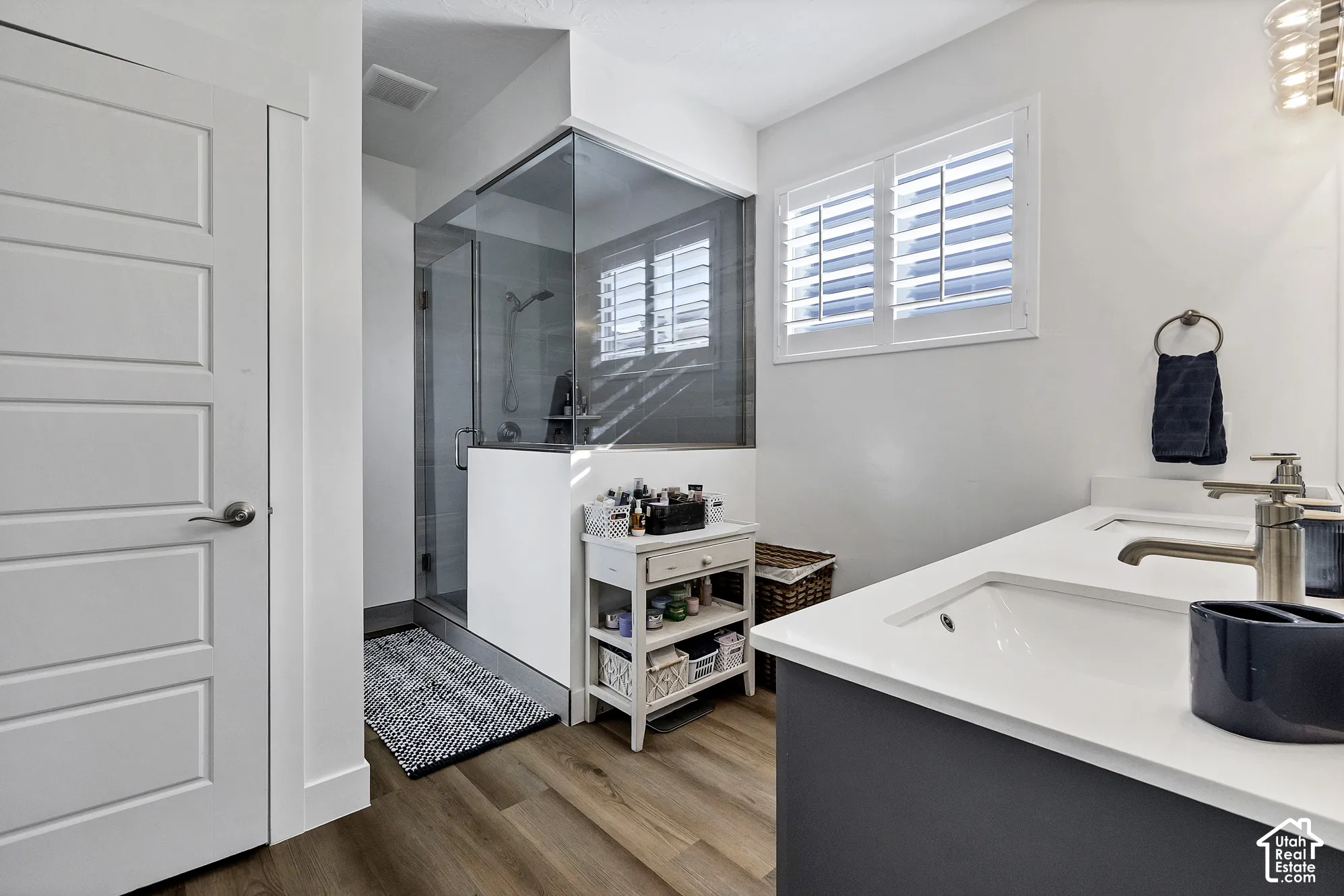Dual vanity with undermount sinks and brushed nickel fixtures, glass-enclosed shower, wood-finish flooring, and window with white shutters