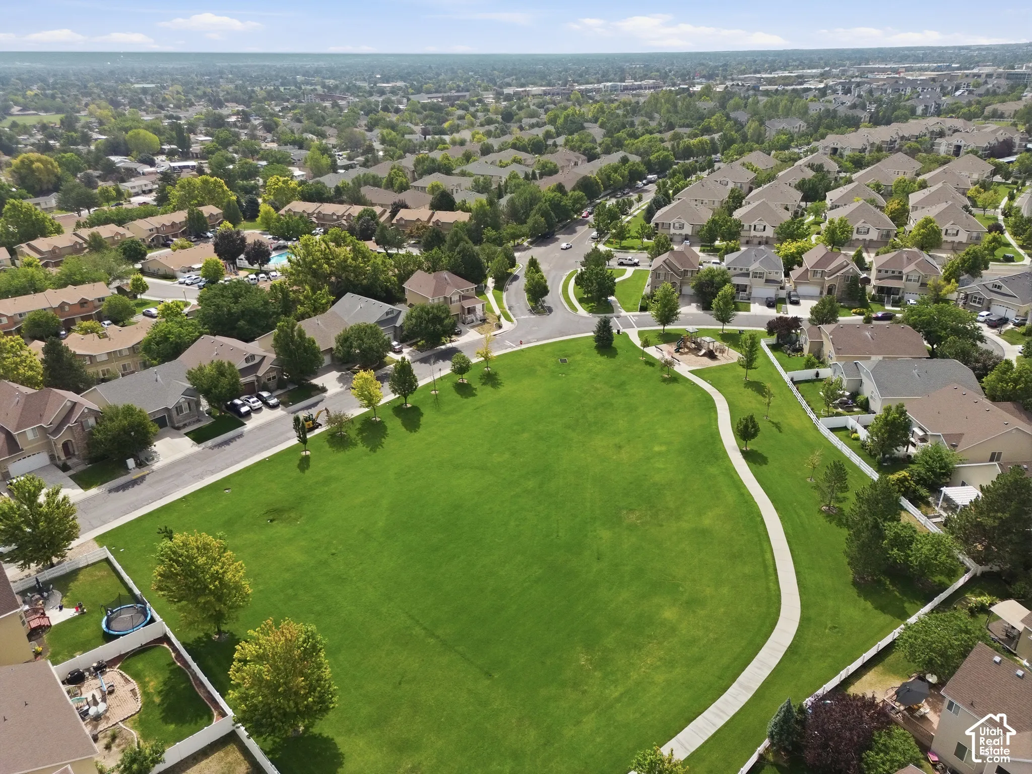 Expansive manicured lawn anchored by mature landscaping and curved hardscape pathways, surrounded by established residential neighborhood with tree-lined streets and single-family homes extending to the horizon