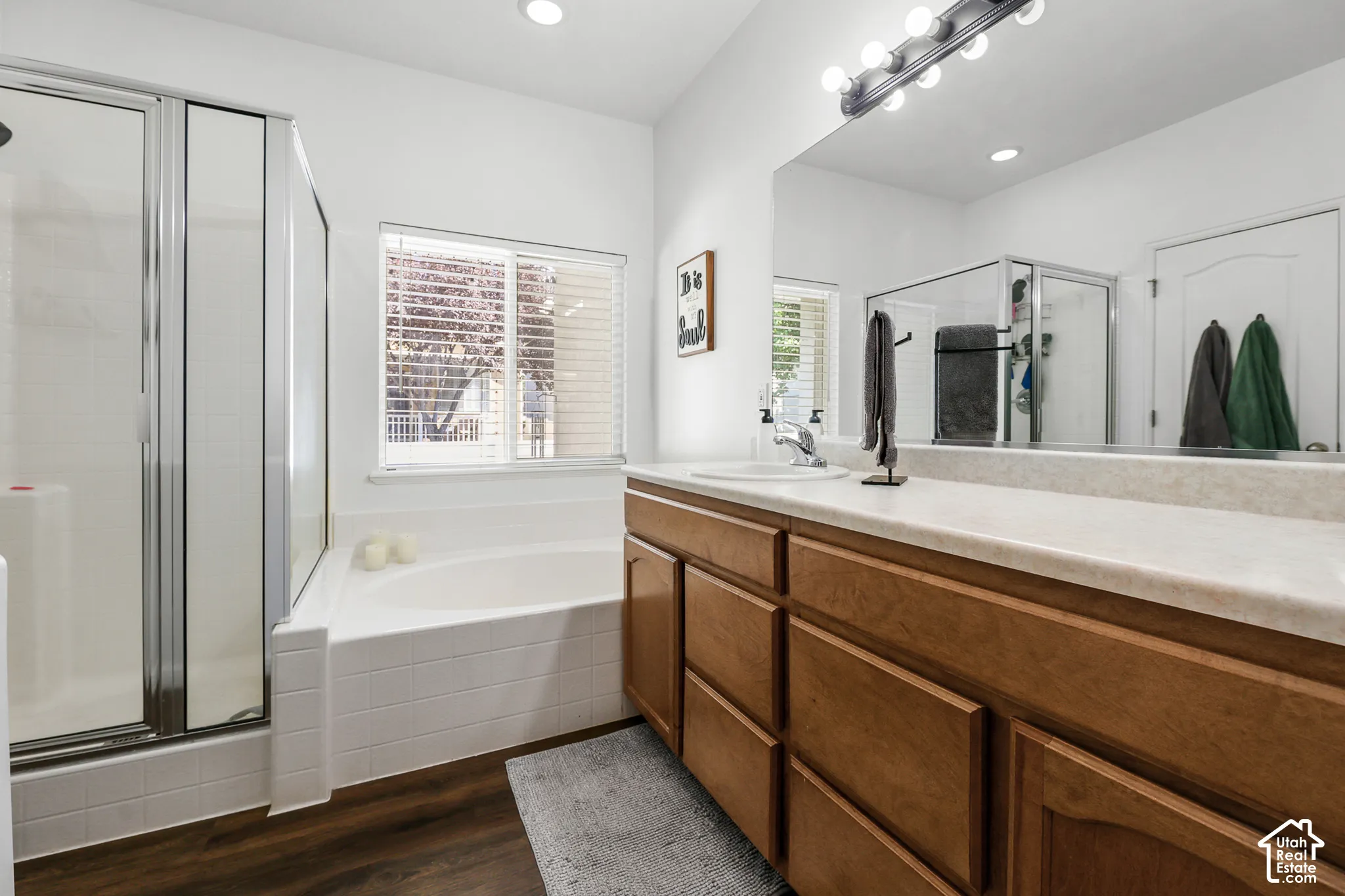 Wood-finish flooring, built-in soaking tub, glass-enclosed shower, wood-finish vanity with dual sinks, and a large mirror with overhead lighting