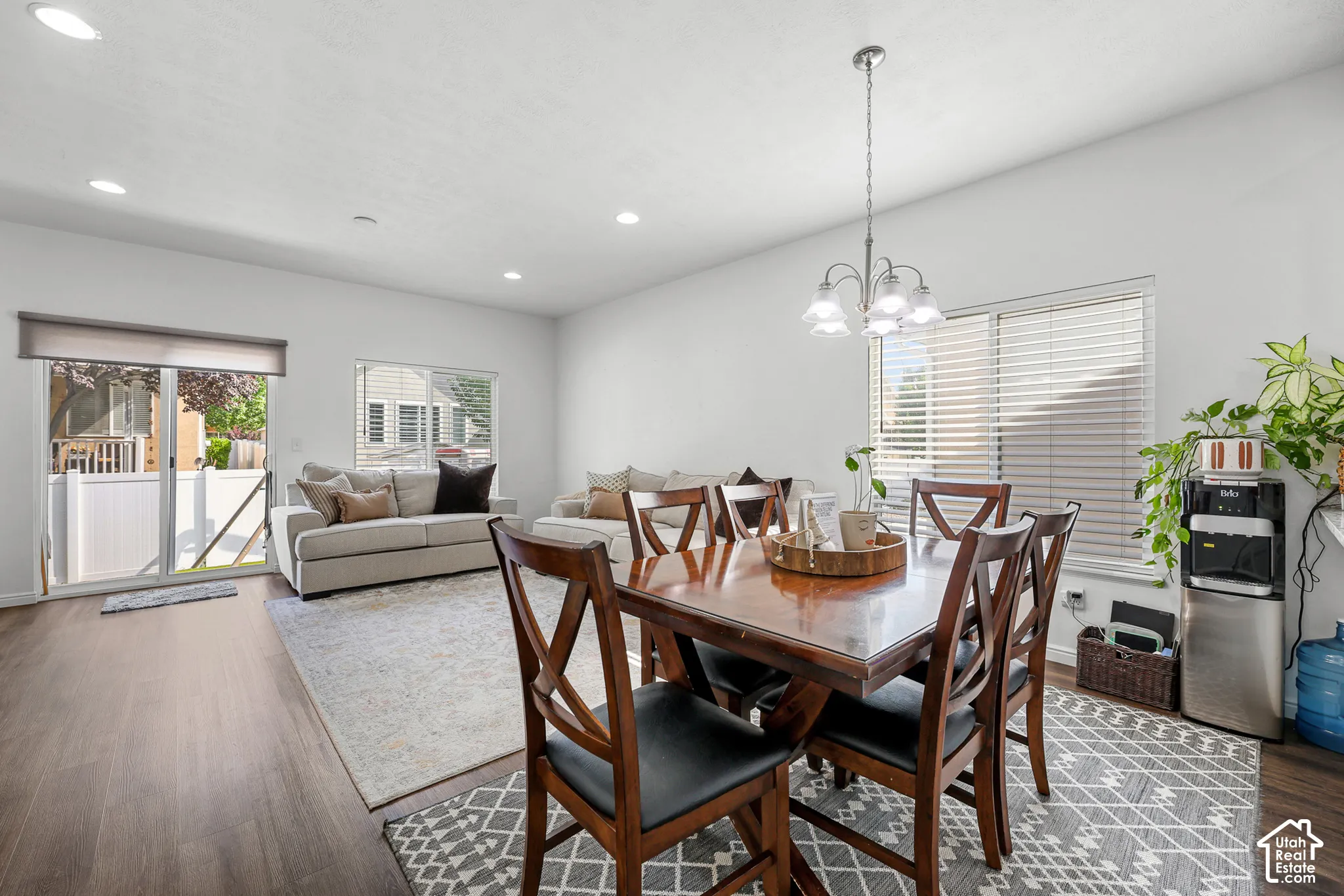 Open-concept dining and living area featuring hardwood flooring, a wood-finish dining table with seating for six, a pendant light fixture with glass shades, and abundant natural light from multiple windows with plantation shutters