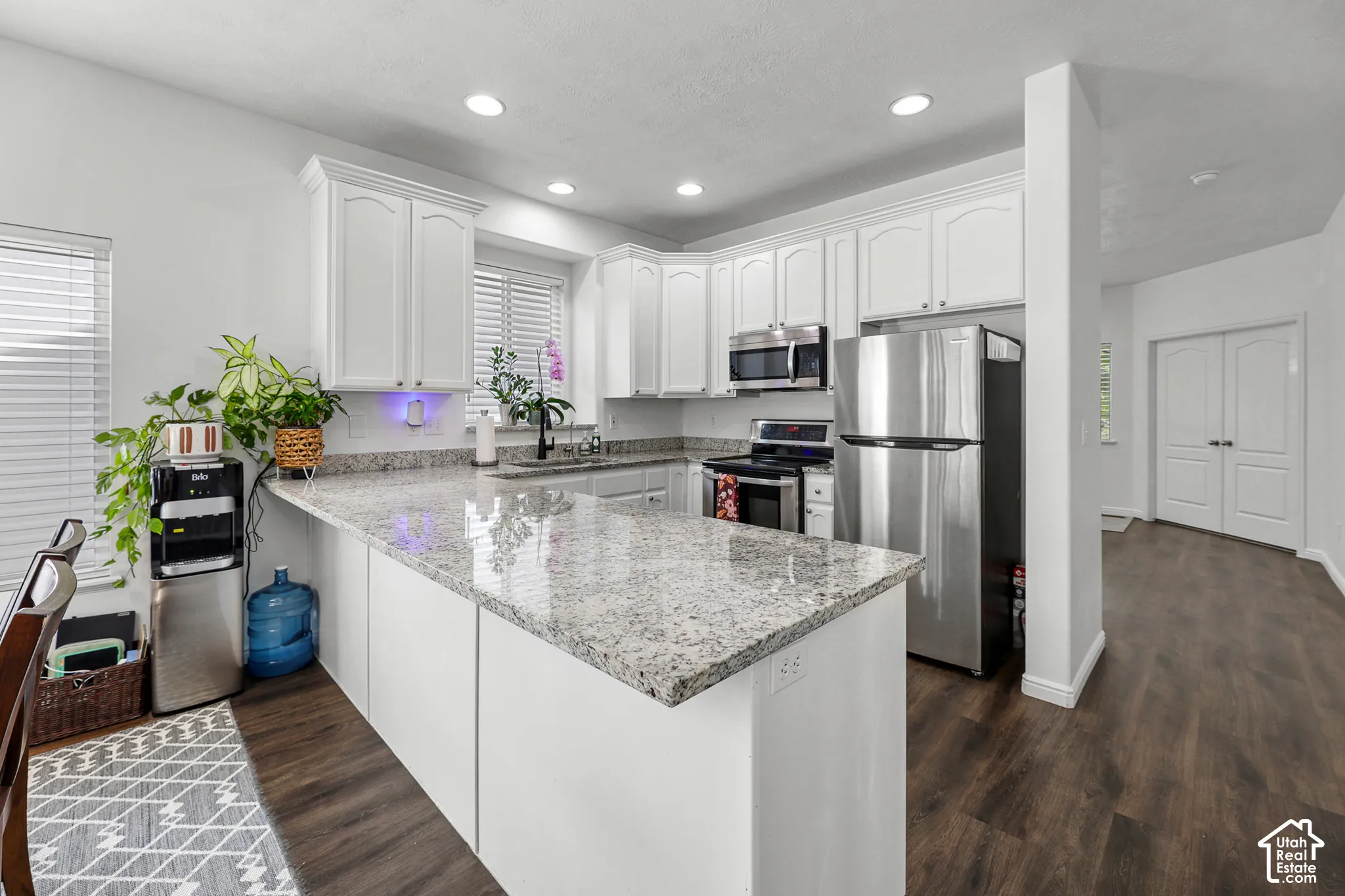 Modern kitchen featuring white cabinetry, stainless steel appliances, granite-style countertops with waterfall island edge, recessed ceiling lighting, and wood-finish flooring throughout the open-concept space