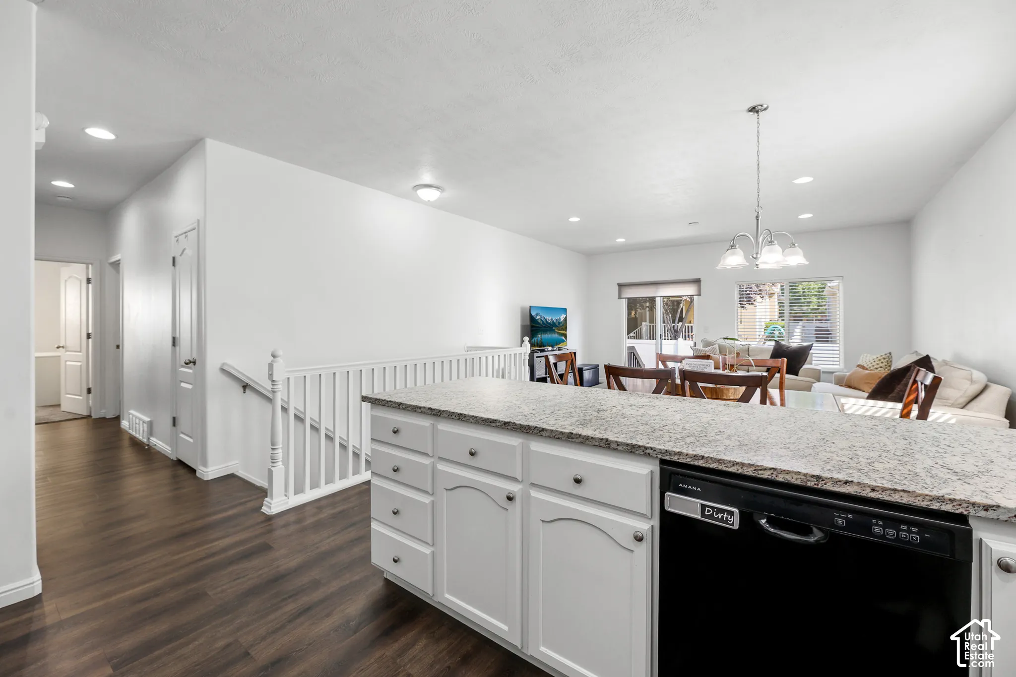 Open-concept kitchen and living area featuring white cabinetry with brushed hardware, quartz countertops, stainless steel appliances including a black dishwasher, wood-finish flooring throughout, recessed and pendant lighting, and a white railed staircase