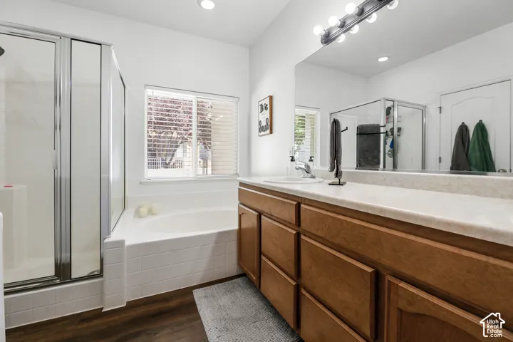 Wood-finish flooring, built-in soaking tub, glass-enclosed shower, wood-finish vanity with dual sinks, and a large mirror with overhead lighting