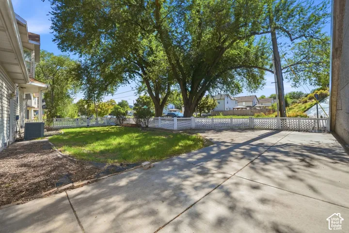 Expansive concrete driveway, manicured lawn, white lattice fencing, mature trees, and a visible exterior HVAC unit