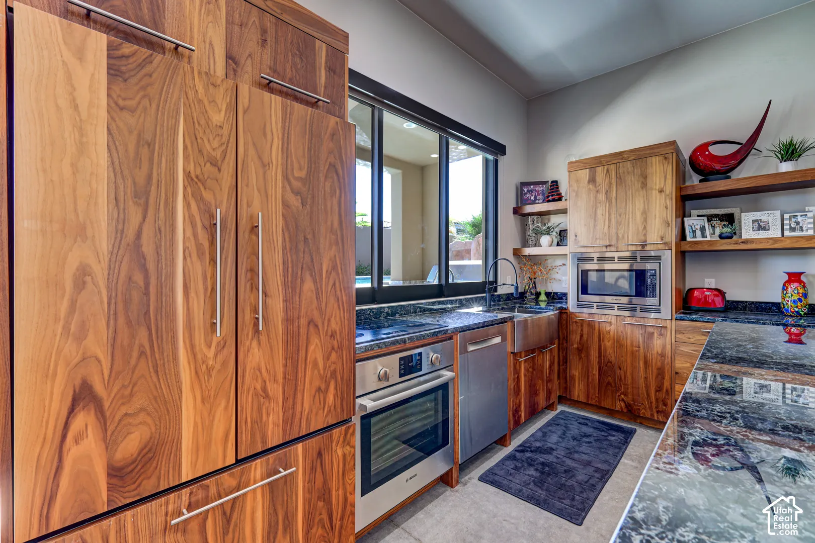 Custom wood-paneled refrigerator, integrated stainless steel oven, large basin sink, dark stone-finish countertops, and wood floating shelves