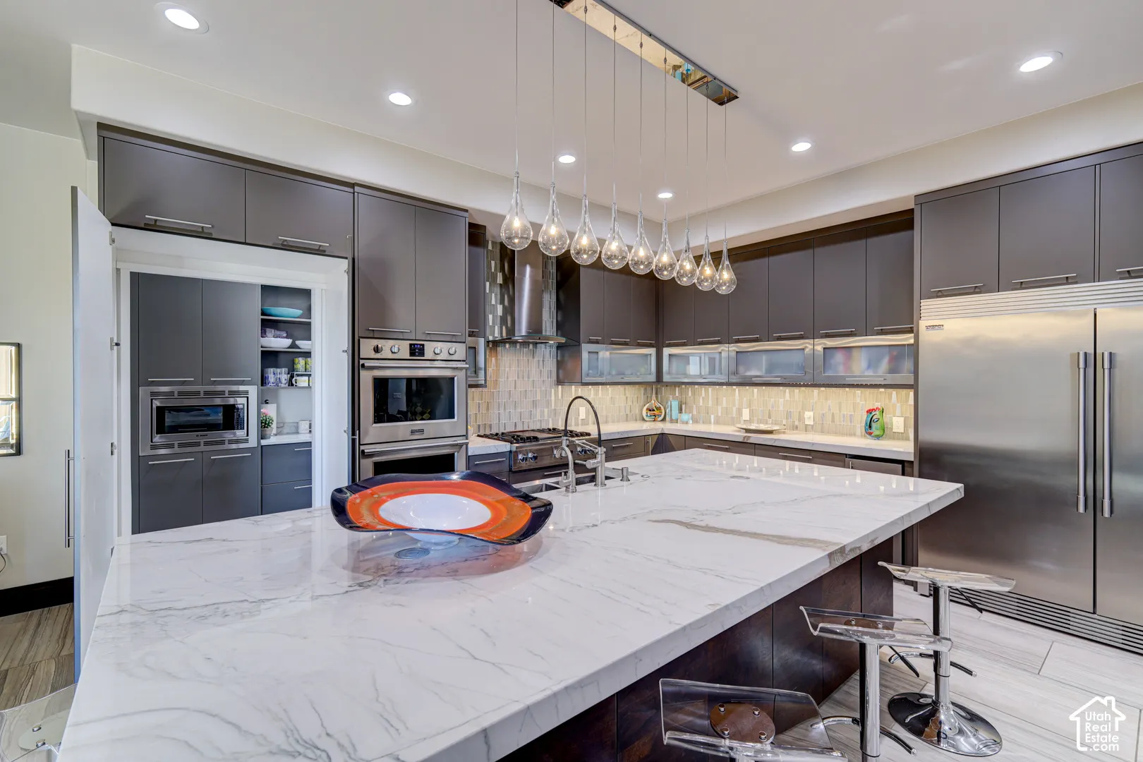 White marble-finish island countertop, stainless steel double oven, stainless steel refrigerator, dark wood-finish cabinetry, and contemporary pendant lighting