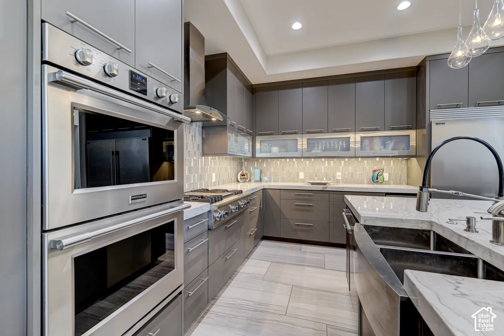 Integrated stainless steel double ovens, gray flat-panel cabinetry, light-toned backsplash, stainless steel range hood, and a double basin farmhouse sink