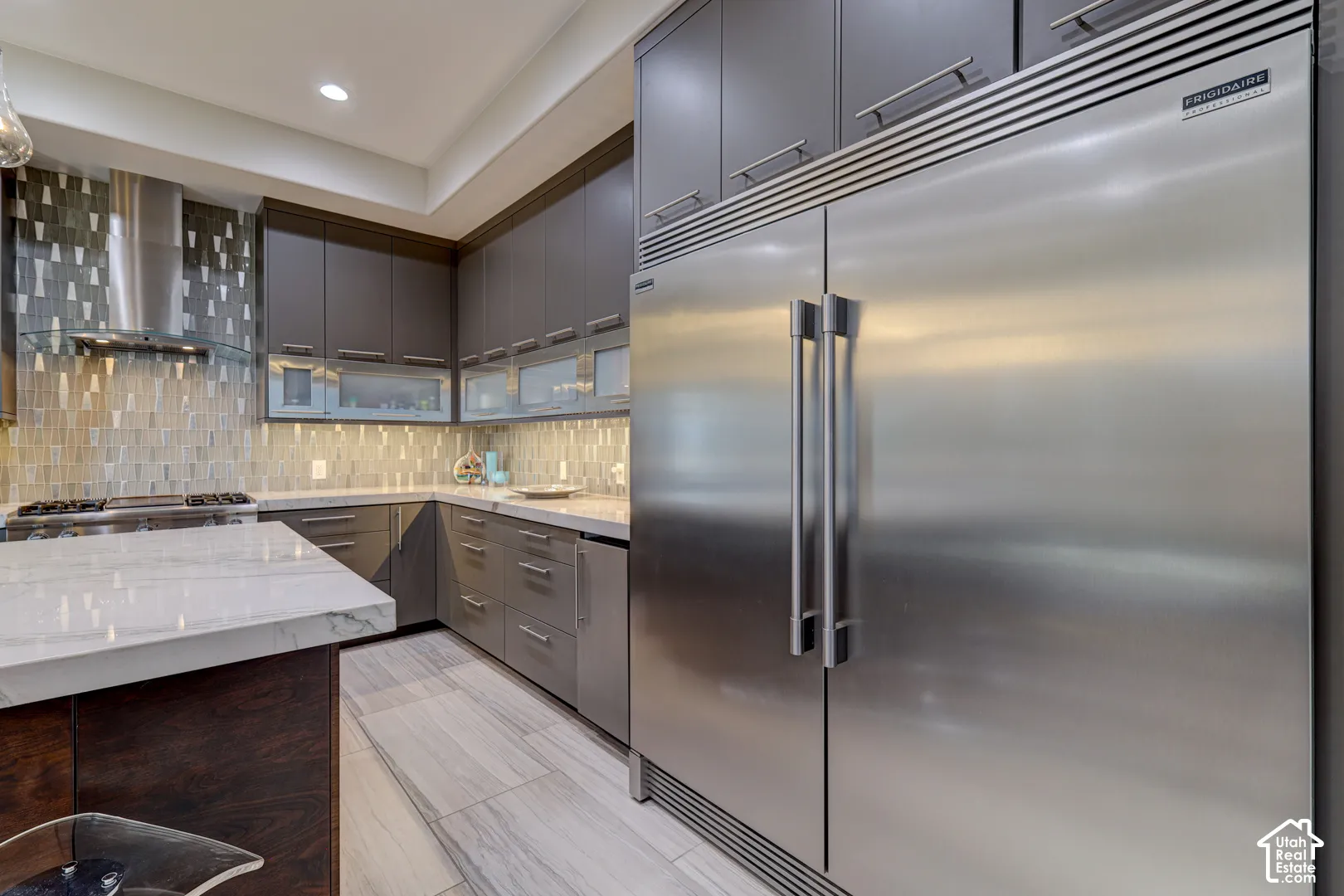 Stainless steel refrigerator with a built-in appearance, gray cabinetry with modern hardware, light-toned stone-finish flooring, and a stone backsplash with varied tile patterns