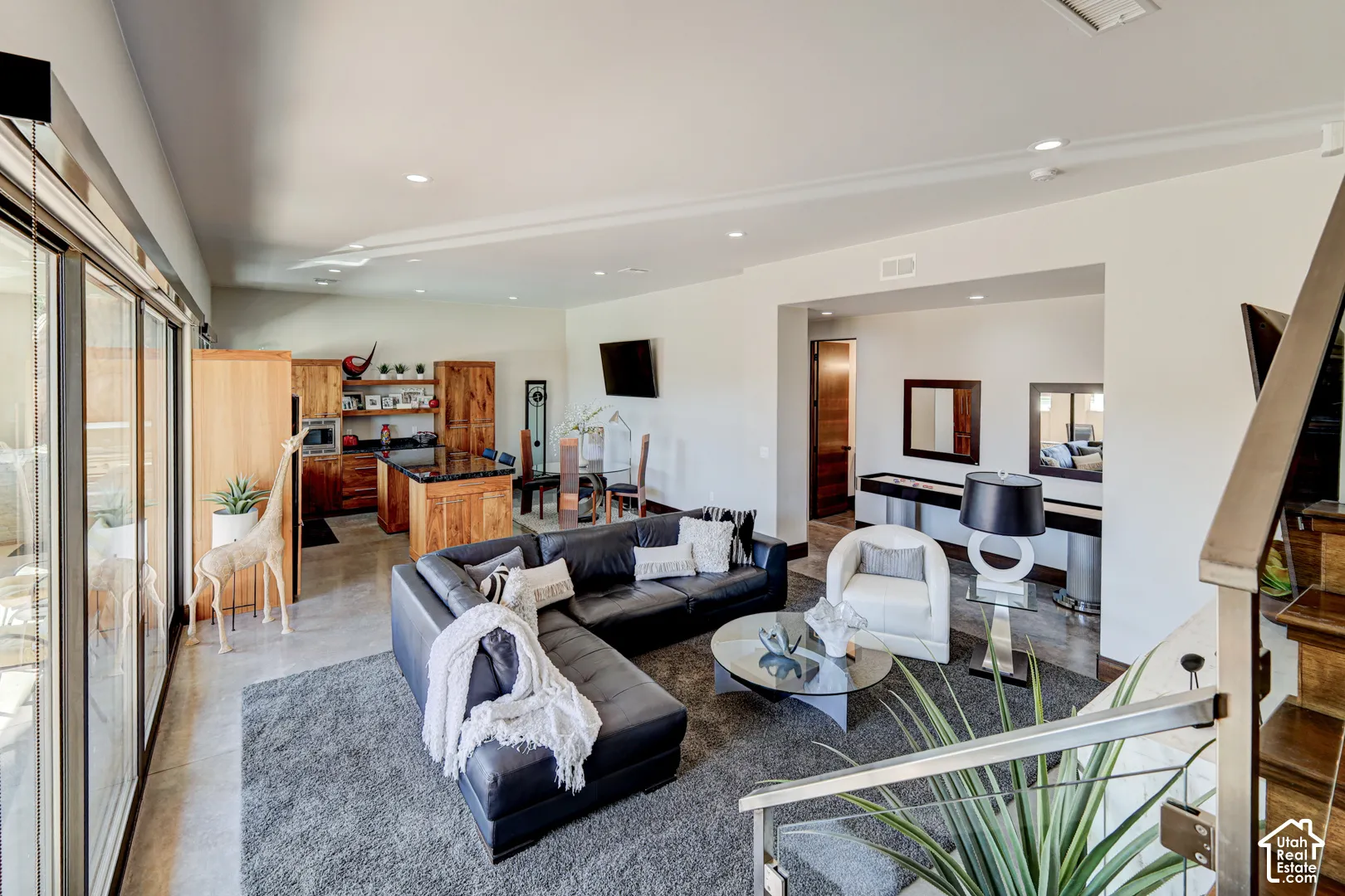 Open-concept living area featuring a kitchen with wood cabinetry and black countertops, concrete-style flooring, a recessed lighting, and a modern stair railing with stainless steel details