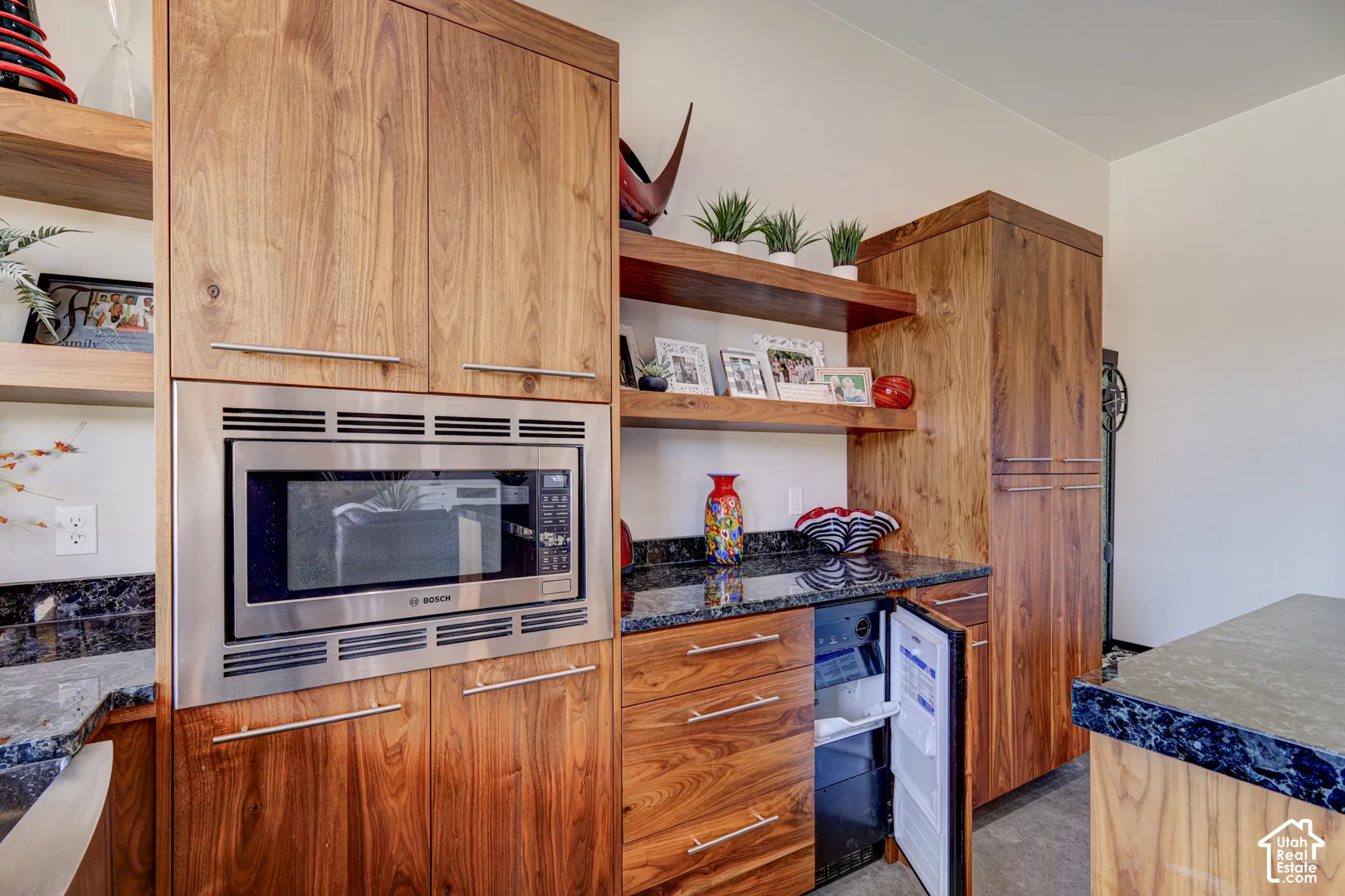 Built-in microwave, undercounter refrigerator, wood-finish cabinetry, dark stone countertops, and floating wood-finish shelving