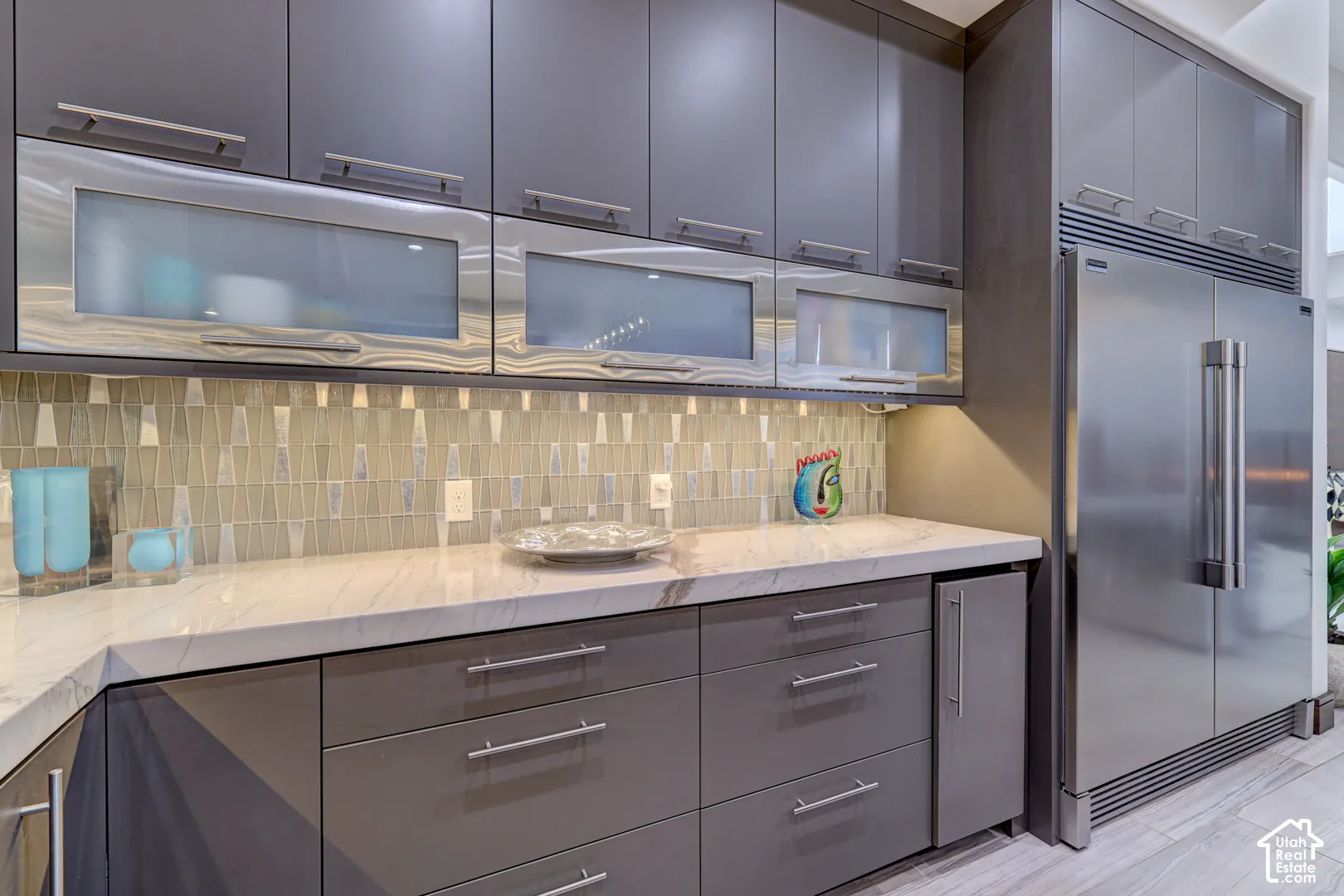 Integrated stainless steel refrigerator, contemporary gray cabinetry, light-toned countertop, mosaic tile backsplash, and under-cabinet lighting
