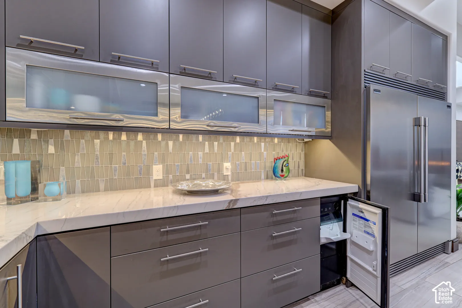 White countertop with gray veining, gray flat-panel cabinetry, stainless steel appliances, glass-front upper cabinets, and a mosaic tile backsplash