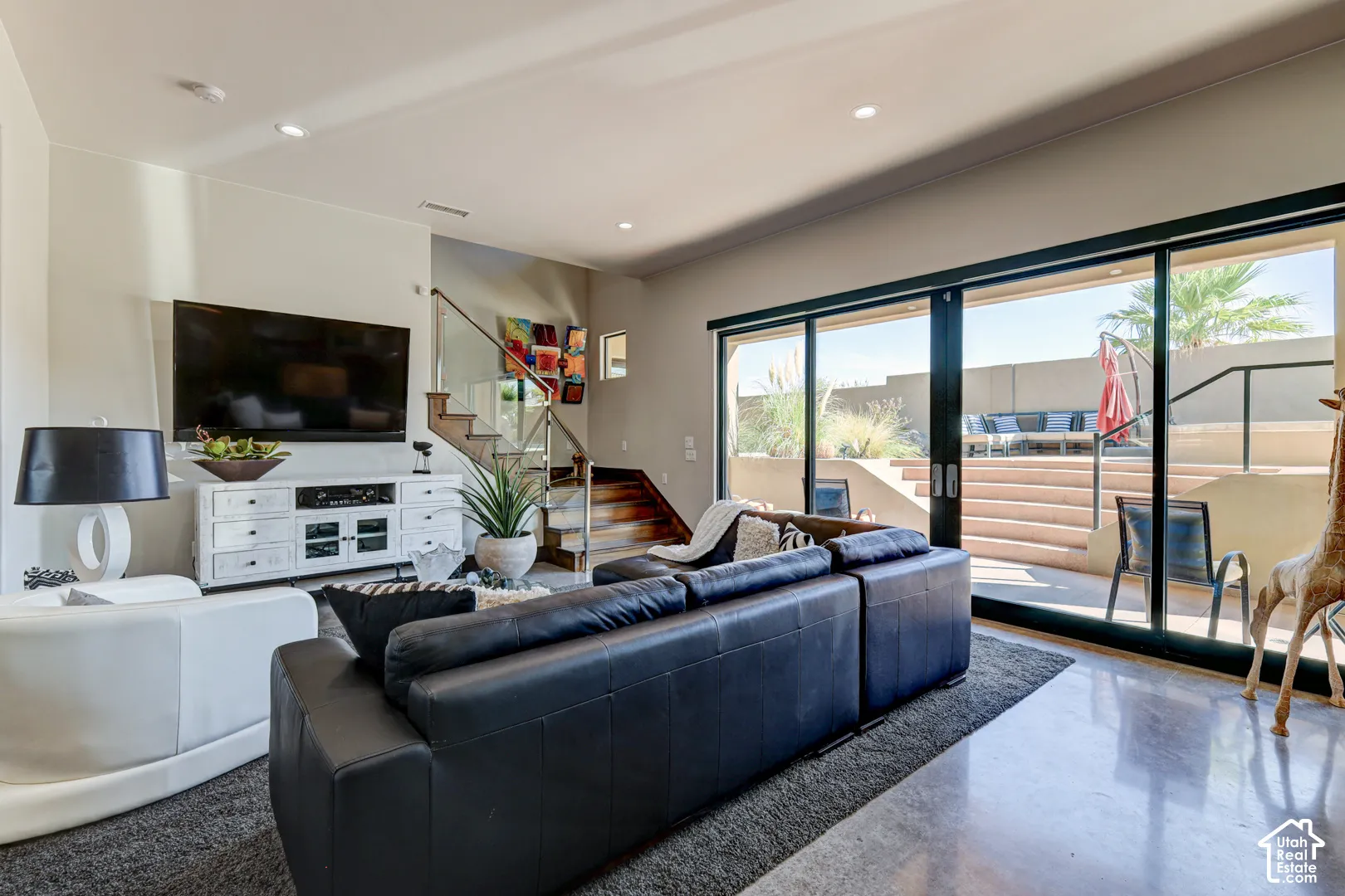 Living area with polished concrete flooring, recessed lighting, a modern staircase with glass railing, and large sliding glass doors leading to an outdoor patio with tiered hardscaping