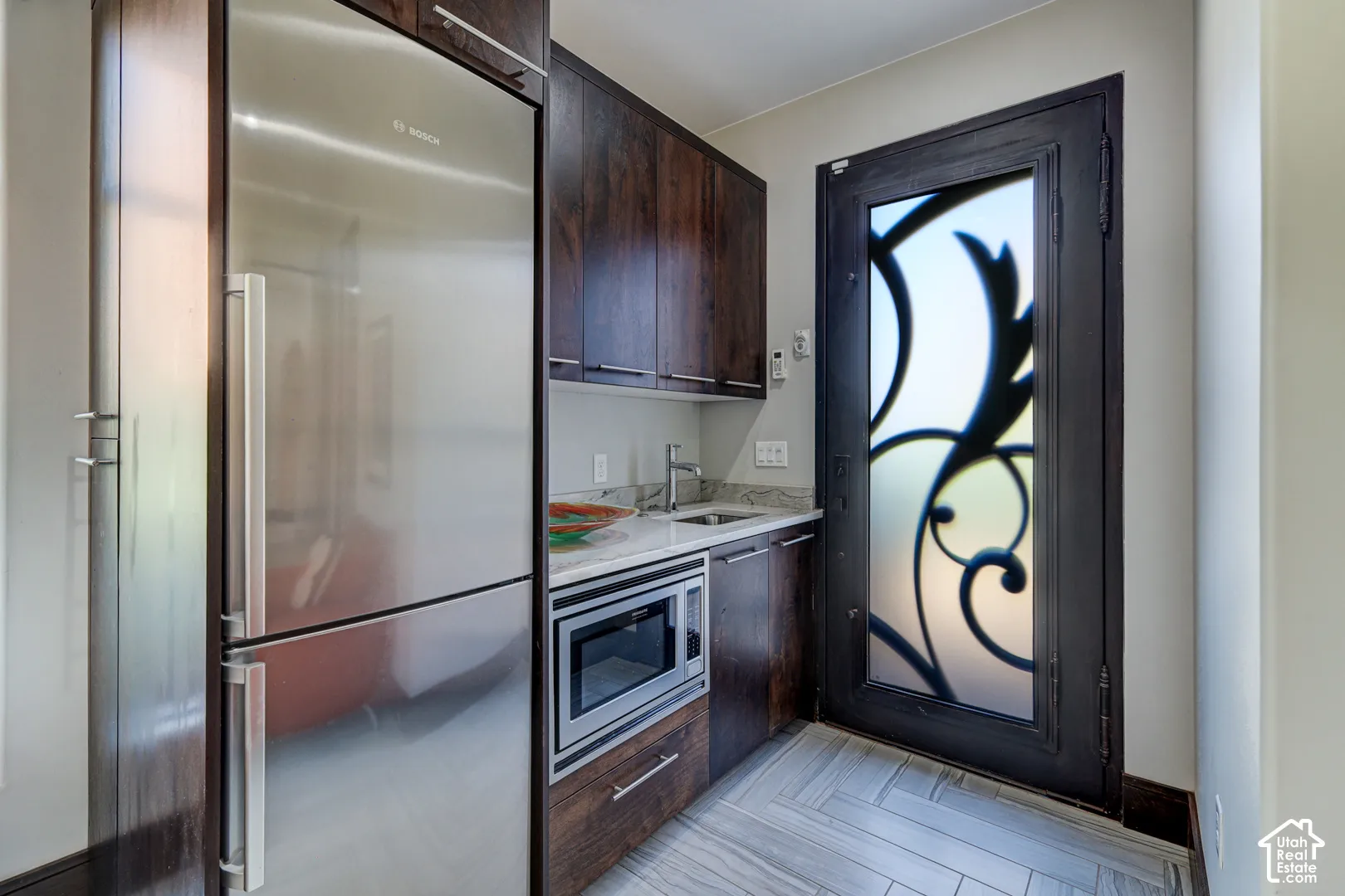 Stainless steel refrigerator and microwave, wood-finish cabinetry, stone countertop, decorative frosted glass door, and herringbone-patterned tile flooring