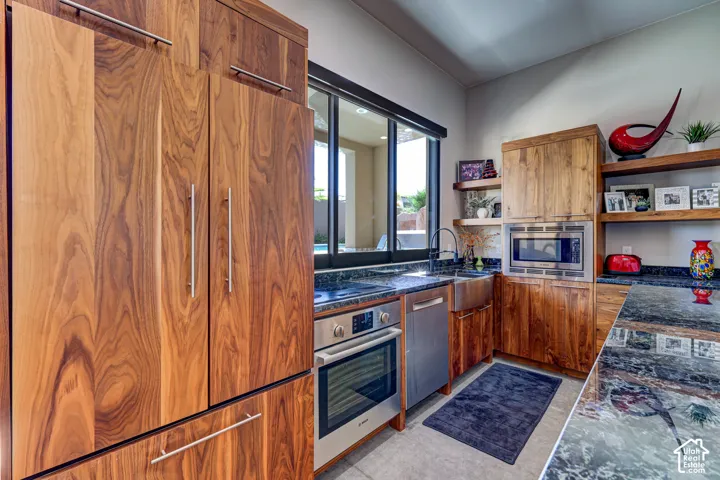 Custom wood-paneled refrigerator, integrated stainless steel oven, large basin sink, dark stone-finish countertops, and wood floating shelves
