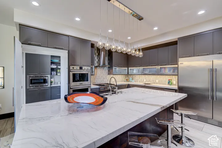 White marble-finish island countertop, stainless steel double oven, stainless steel refrigerator, dark wood-finish cabinetry, and contemporary pendant lighting