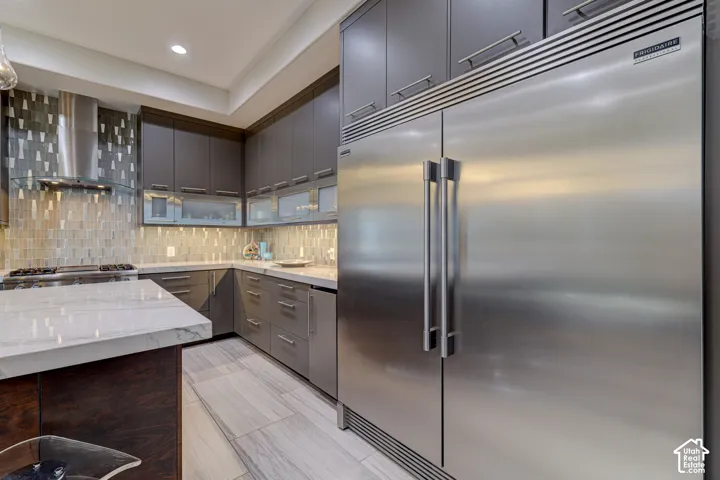 Stainless steel refrigerator with a built-in appearance, gray cabinetry with modern hardware, light-toned stone-finish flooring, and a stone backsplash with varied tile patterns