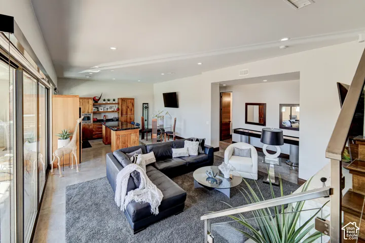 Open-concept living area featuring a kitchen with wood cabinetry and black countertops, concrete-style flooring, a recessed lighting, and a modern stair railing with stainless steel details